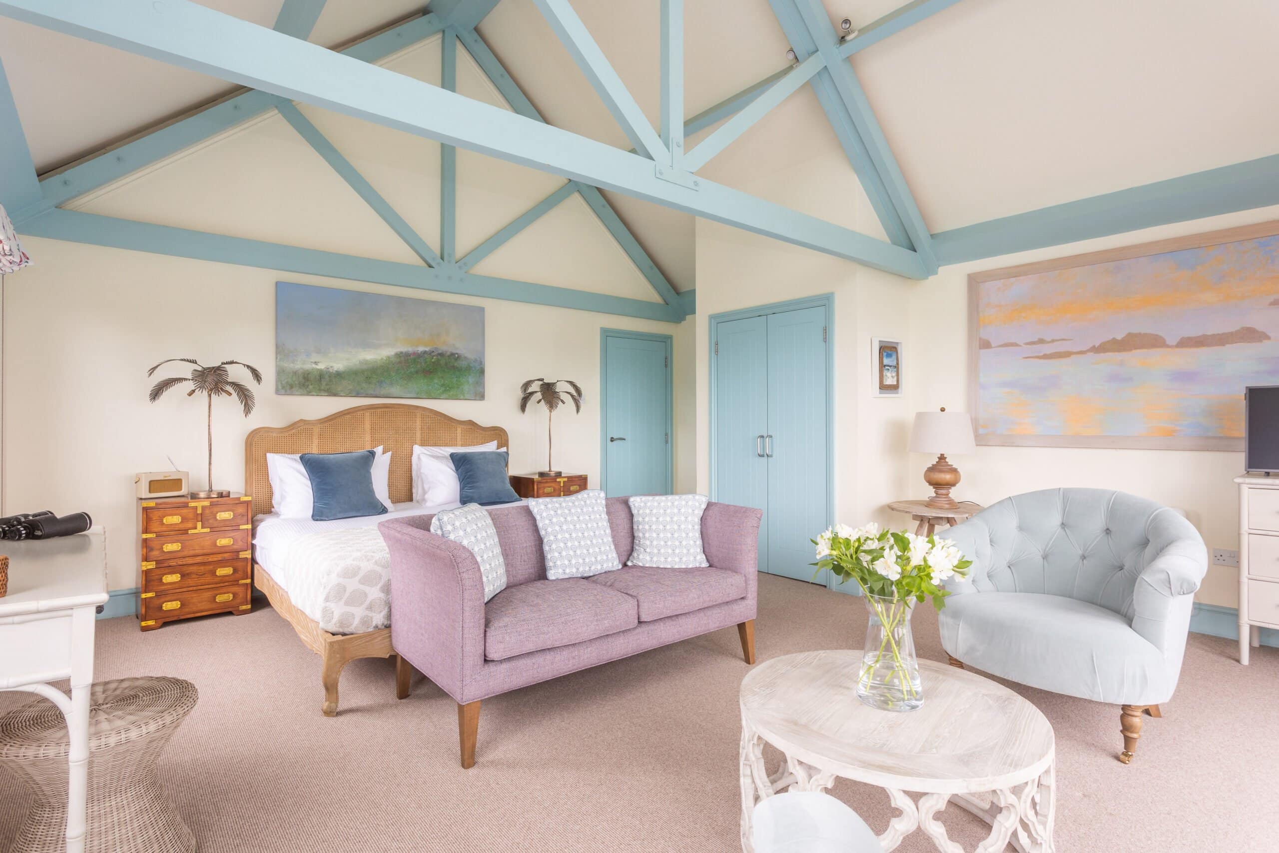 A spacious bedroom with a wooden bed, white and blue bedding, a lilac sofa, and a round table with flowers. The room has light blue exposed beams, pastel walls, and landscape paintings, creating a cosy and airy feel.