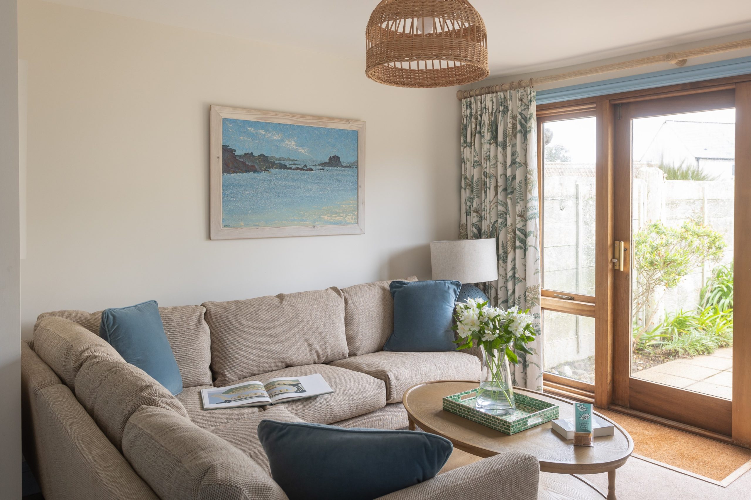 A cosy living room with a beige sectional sofa adorned with blue cushions. There's a round wooden coffee table, a woven pendant light, and a painting on the wall. A glass door with floral curtains opens to a garden view.
