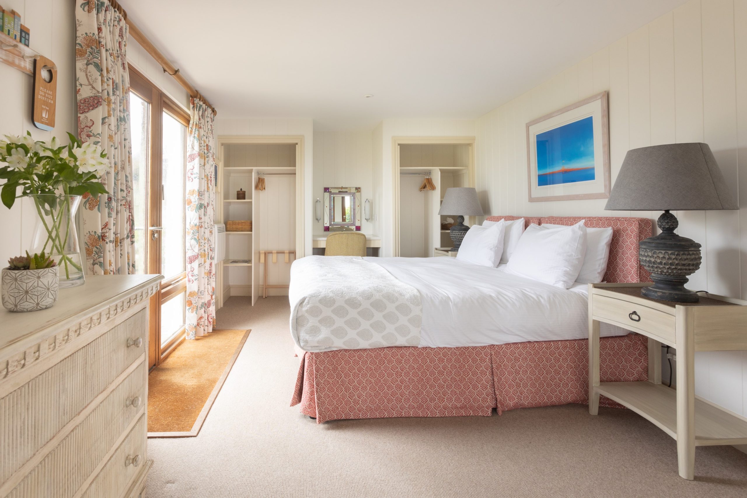 A cosy hotel room with a large bed, white and pink bedding, and two bedside tables with lamps. Floral curtains frame a sliding glass door leading to a balcony. A dresser with flowers and a mirror is against the wall. The décor is light and airy.