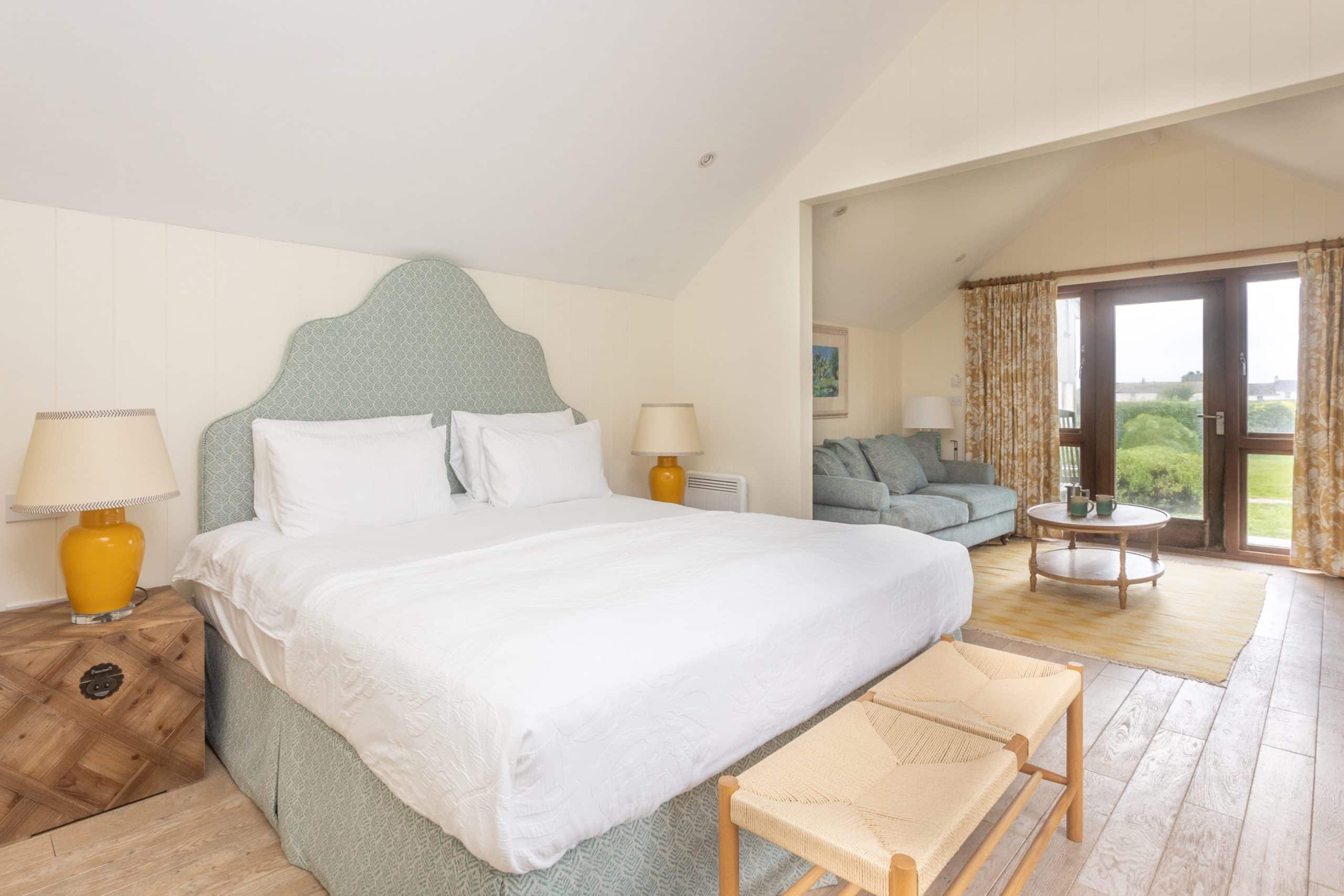 Bright, spacious bedroom with a large bed, pastel green headboard, and white bedding. Two wooden lamps on nightstands flank the bed. A sitting area with a sofa opens to a balcony through glass doors. Light wood flooring and soft natural light enhance the room.