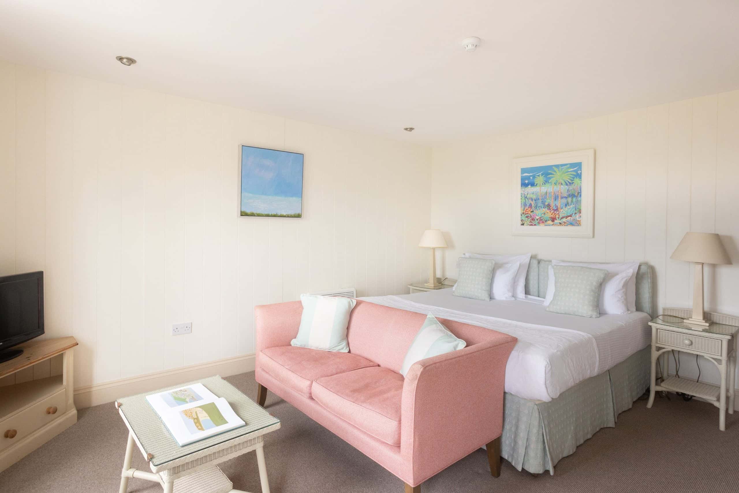 A bright hotel room features a double bed with white linens and pastel pillows, a pink sofa, and light wooden furniture. Two framed artworks are on the walls, and a TV is placed on a corner table. A tray with items rests on the coffee table.