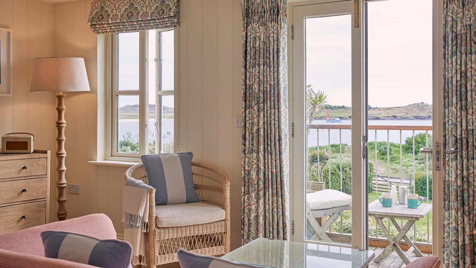 Cosy room with wicker chairs and soft blue and pink tones. A glass coffee table sits atop a patterned rug. Large windows and a glass door reveal a view of greenery and the sea outside. A tall lamp and wooden drawers complete the setting.