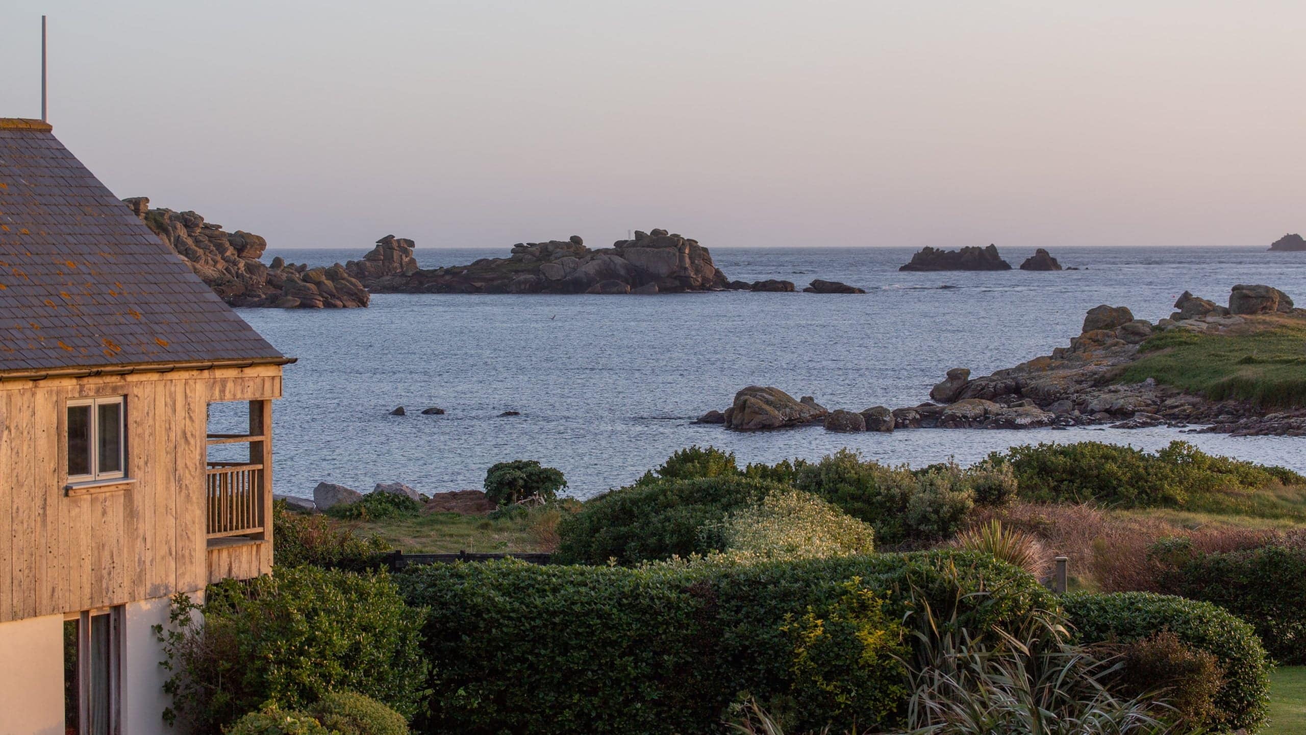 A coastal scene at sunset with the wood-clad Juno bedroom building and balcony partially visible on the left. The foreground features lush green bushes, while rocky formations and calm sea waters extend into the distance under a soft, pastel sky.
