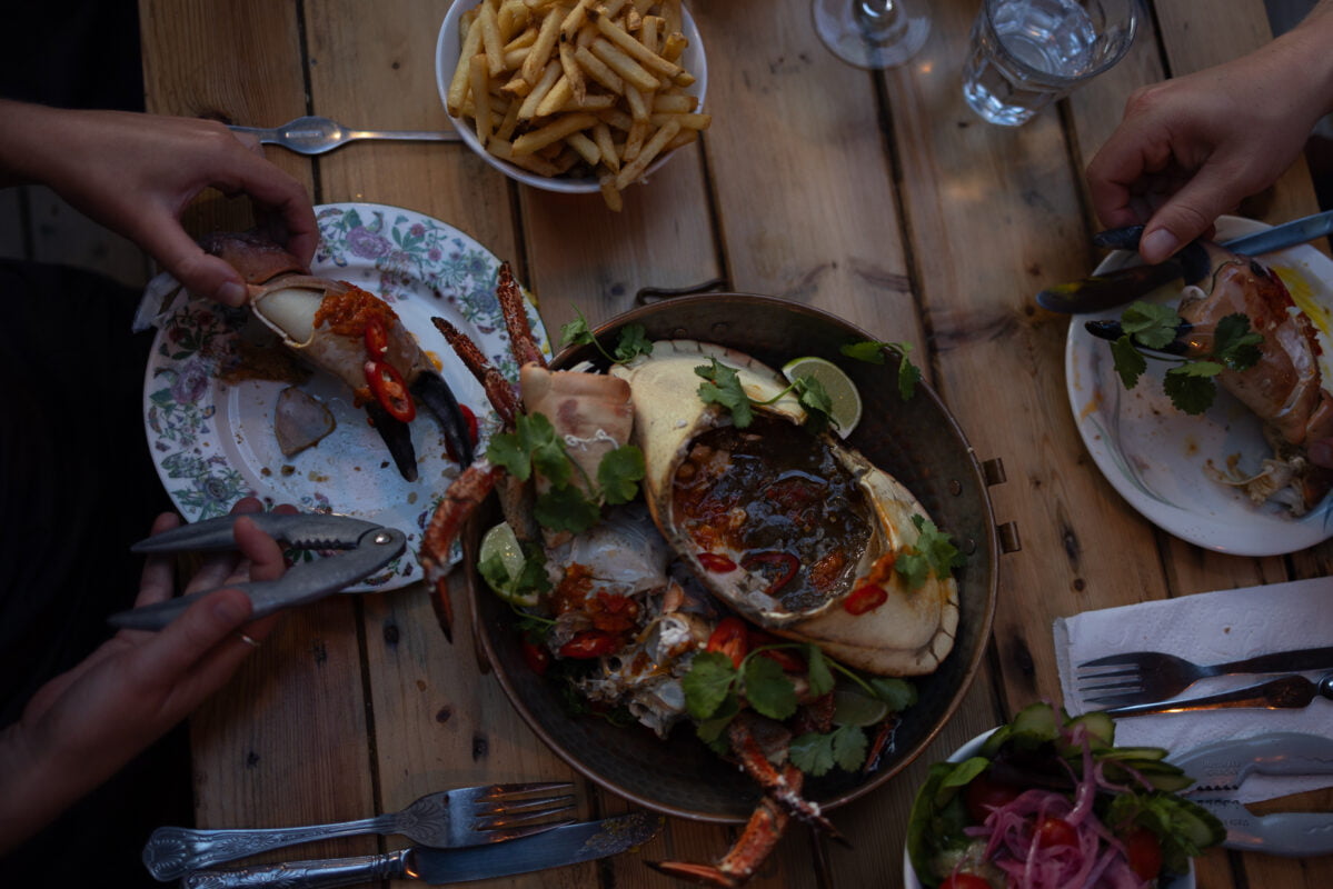 Top view of a table with a seafood platter featuring a large crab garnished with herbs and lime slices. Plates with crab claws, a bowl of fries, and salads are around. Diners are using utensils to serve themselves.