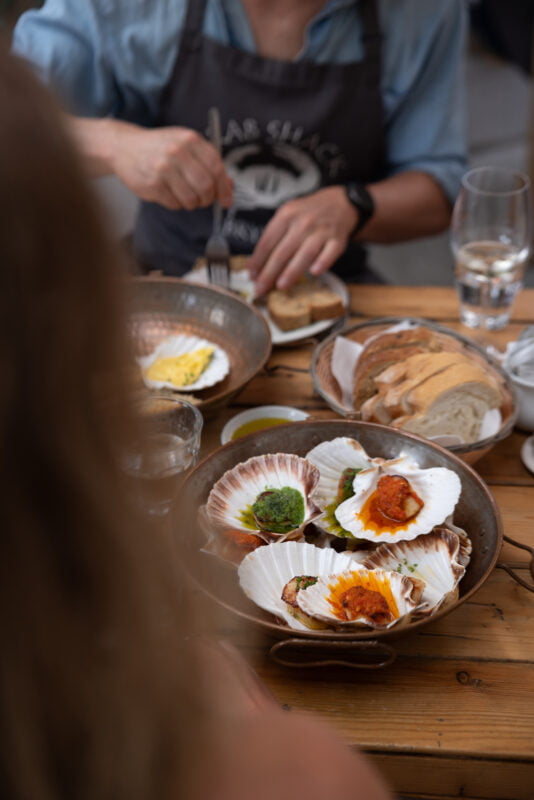 A person is seated at a wooden table with several plates of food, including scallops on shells with various toppings, sliced bread, and dipping sauces. The focus is on the scallops, with the person partially visible in the background.