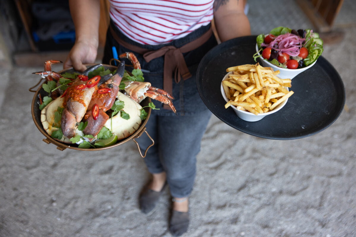 A person wearing a striped shirt and apron is carrying two plates of food. One plate has a seafood dish with garnish, and the other plate has a bowl of french fries and a fresh salad with cherry tomatoes and pickled onions.