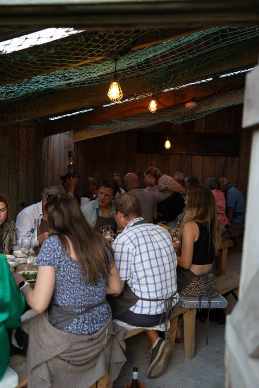 A group of people gather at an outdoor, rustic wooden dining space with soft lighting. Tables are set with food and drinks. Some guests engage in conversation, while others eat. Decorative nets and wooden walls create a cozy, intimate atmosphere.