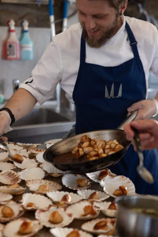 A chef with a blue apron is smiling as he places seared scallops onto a tray of empty shells using a pan. The kitchen setting includes cleaning supplies and a steel countertop. Another hand assists with a spoon.
