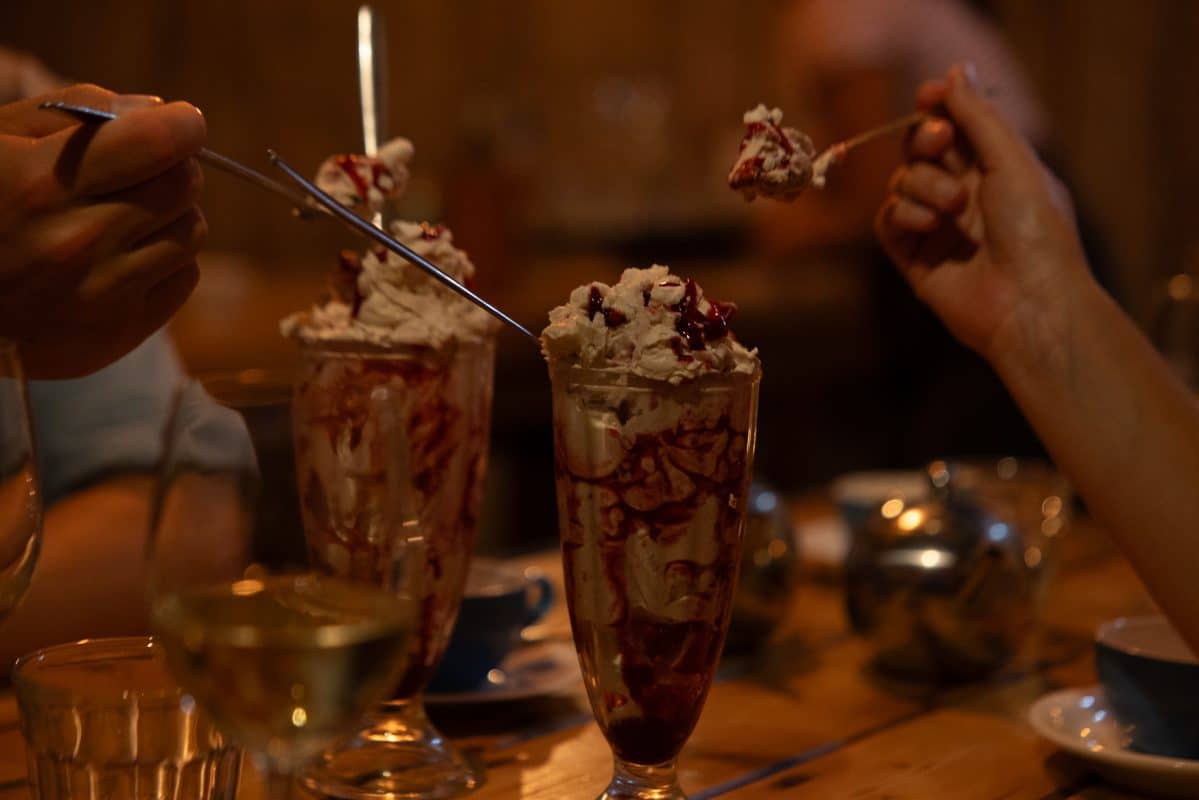 Two people enjoying tall, elegant glasses of sundaes topped with whipped cream and red syrup. The scene is dimly lit, creating a cosy ambiance with additional wine glasses and teacups on the wooden table.
