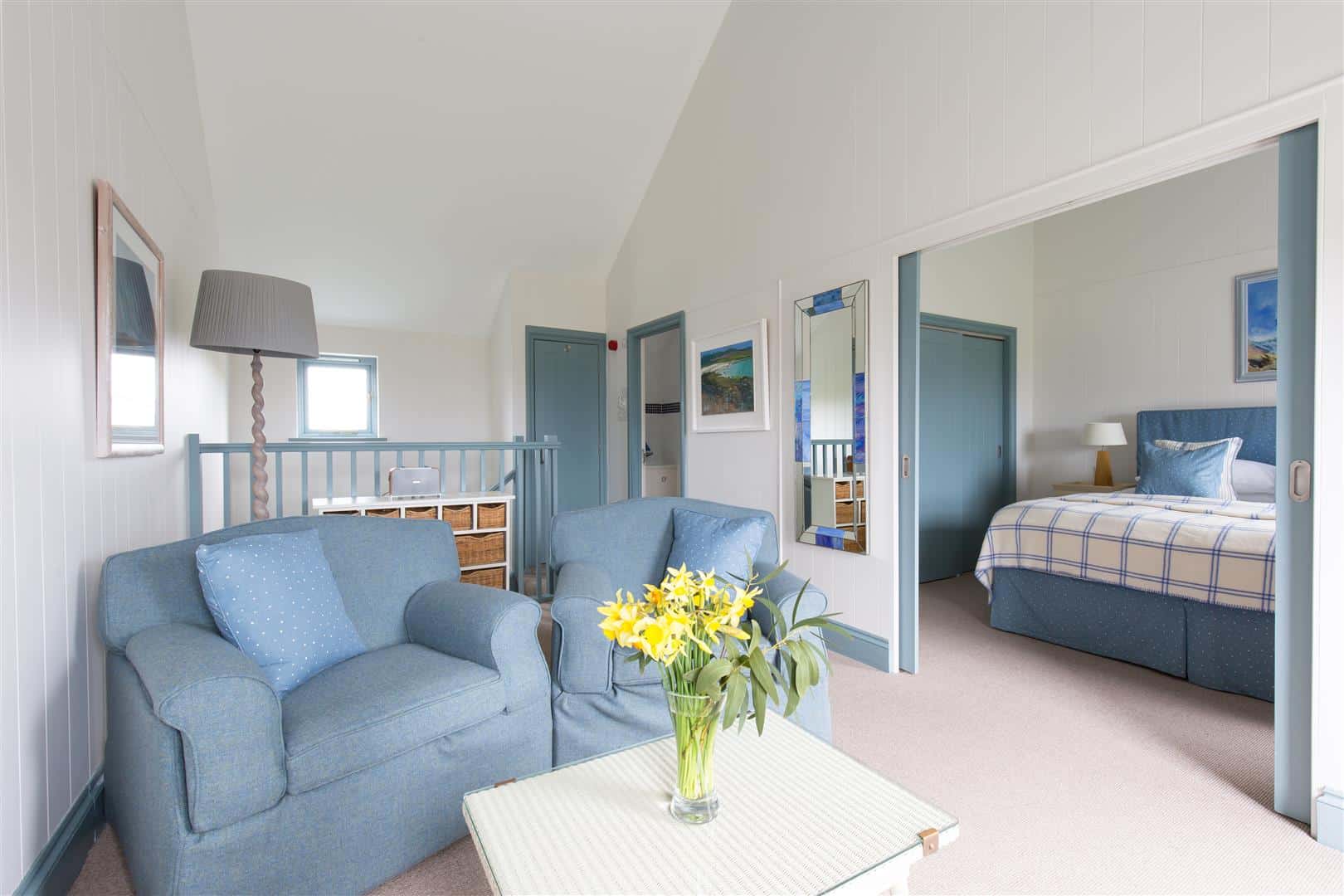 Hotel suite with two large armchairs in a sitting area with fresh flowers, and a separate bedroom with king size bed visible through sliding doors