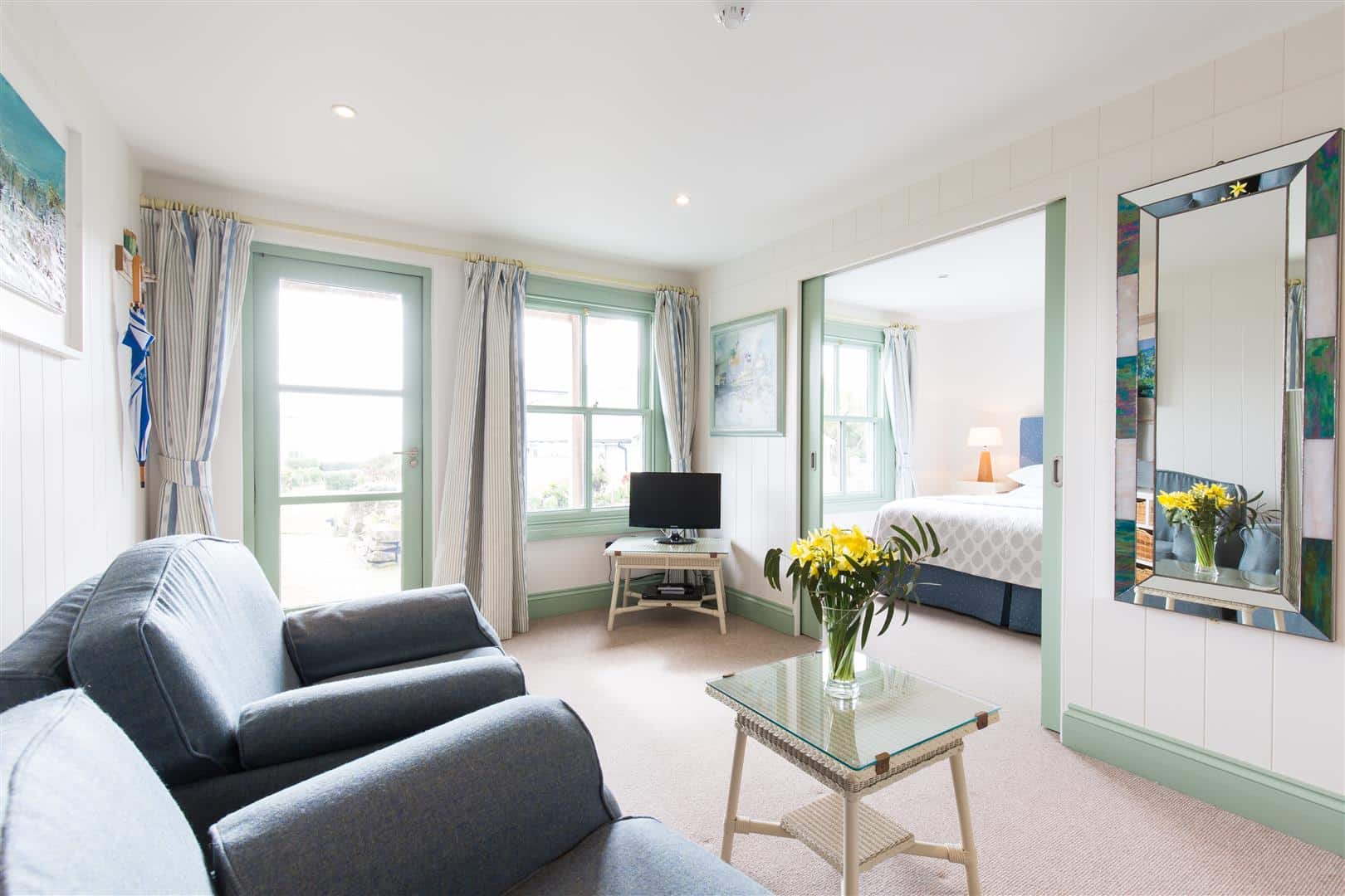 A hotel suite sitting room with two archairs and a television, with a king size bed visible in a separate bedroom through sliding doors