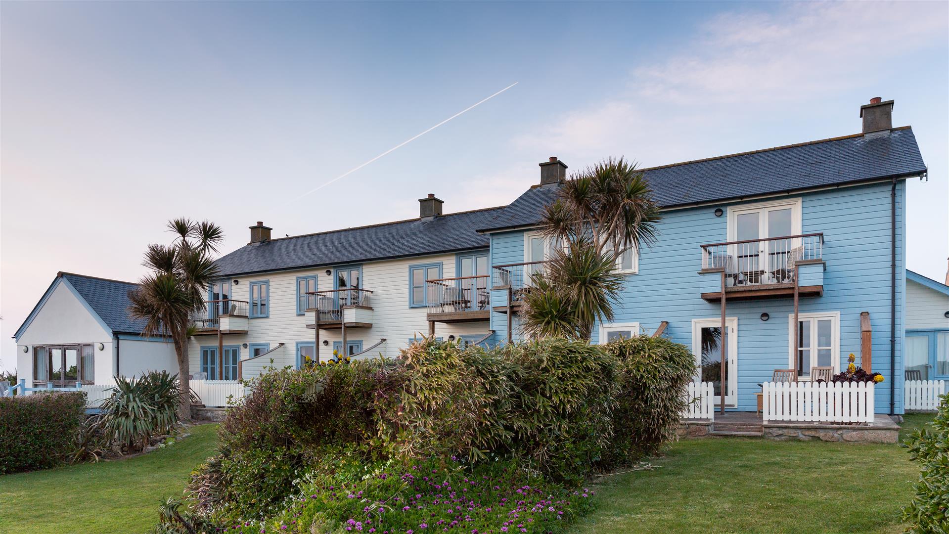 A front view of Hell Bay Hotel with charming blue and white coastal houses with balconies, surrounded by lush greenery and palm trees. A clear sky adds to the serene atmosphere.