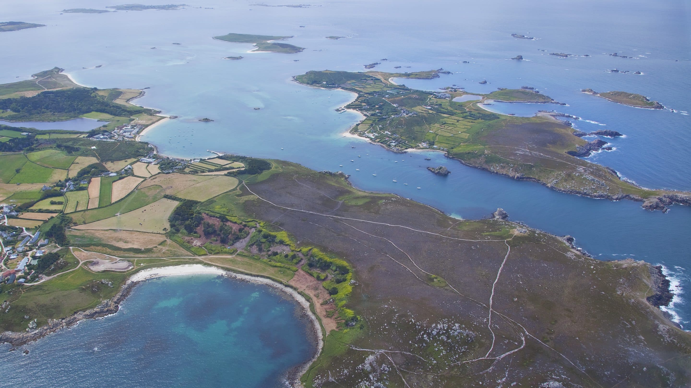 Tresco and Bryher North End Aerial view of a coastline with a mix of farmland, rocky terrain, and small beaches surrounded by blue ocean, with several smaller islands scattered offshore.